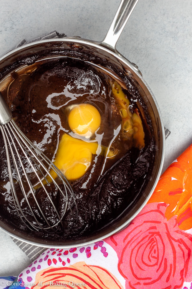 brownie batter in a silver pan with eggs being added with a wire whisk on a light white/ grey surface with a colorful multi colored linen