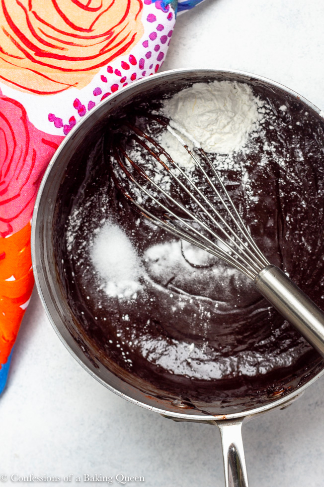 baking soda, cornstarch and salt being added to brownie batter with a whisk on a light white/ grey surface with a colorful multi colored linen