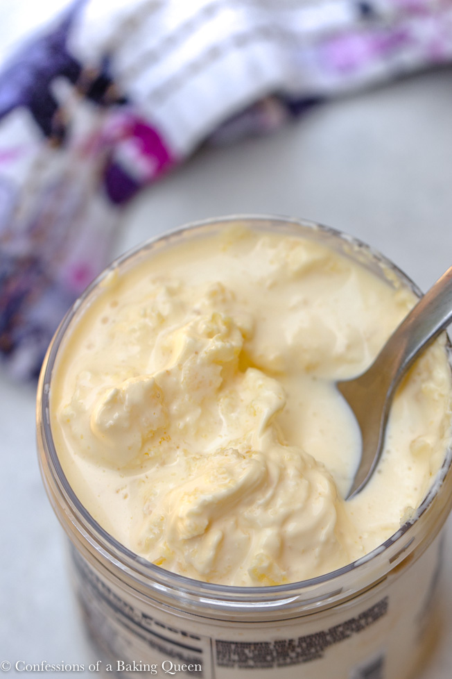 clotted cream in a clear container with a spoon taking a spoonful on a white background next to a purple, white, and pink linen