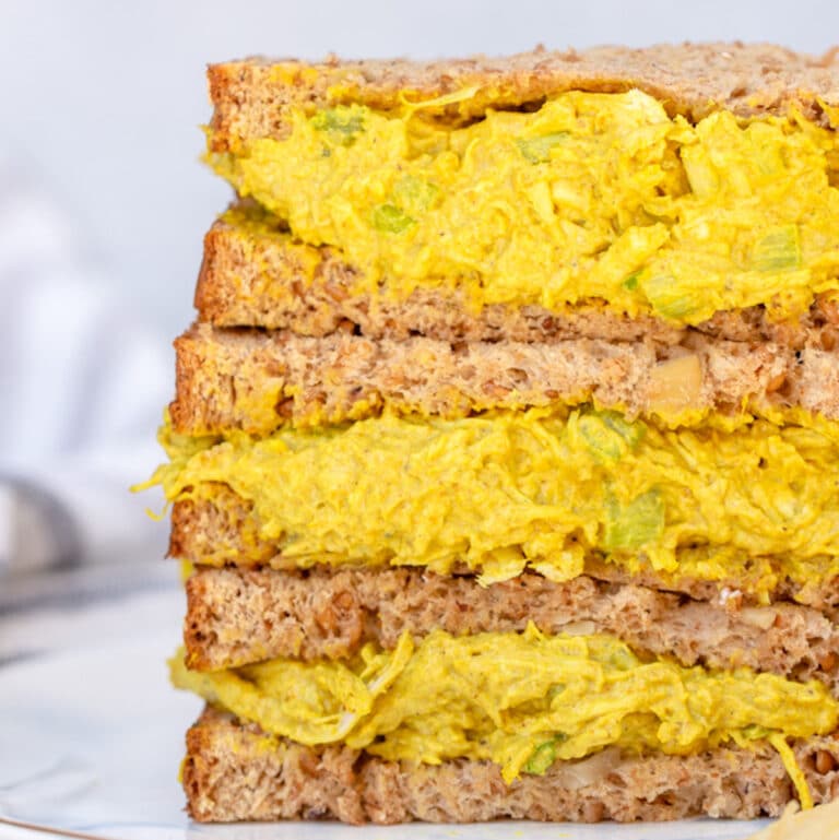 close up of curried chicken salad recipe served in a sandwich, three halves of sandwiches stacked on top of each other on a white and blue plate on a white background