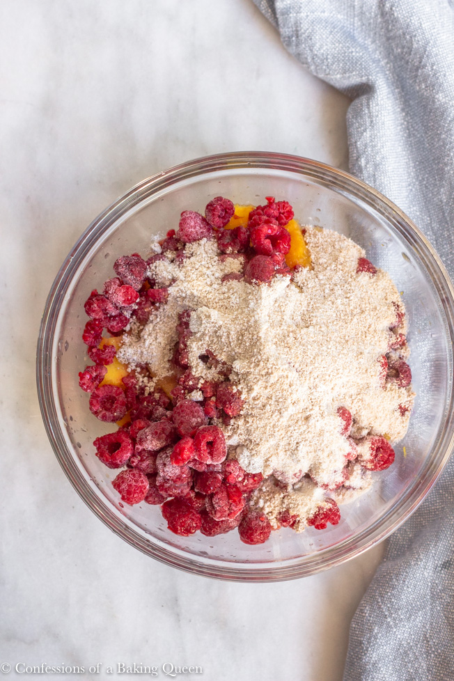 raspberries, sugar, flour, and peaches mixed together in a large clear bowl on a marble surface with a light blue linen