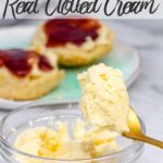 clotted cream in a clear bowl with a gold spoon holding a scoop with scones in the background on a white surface