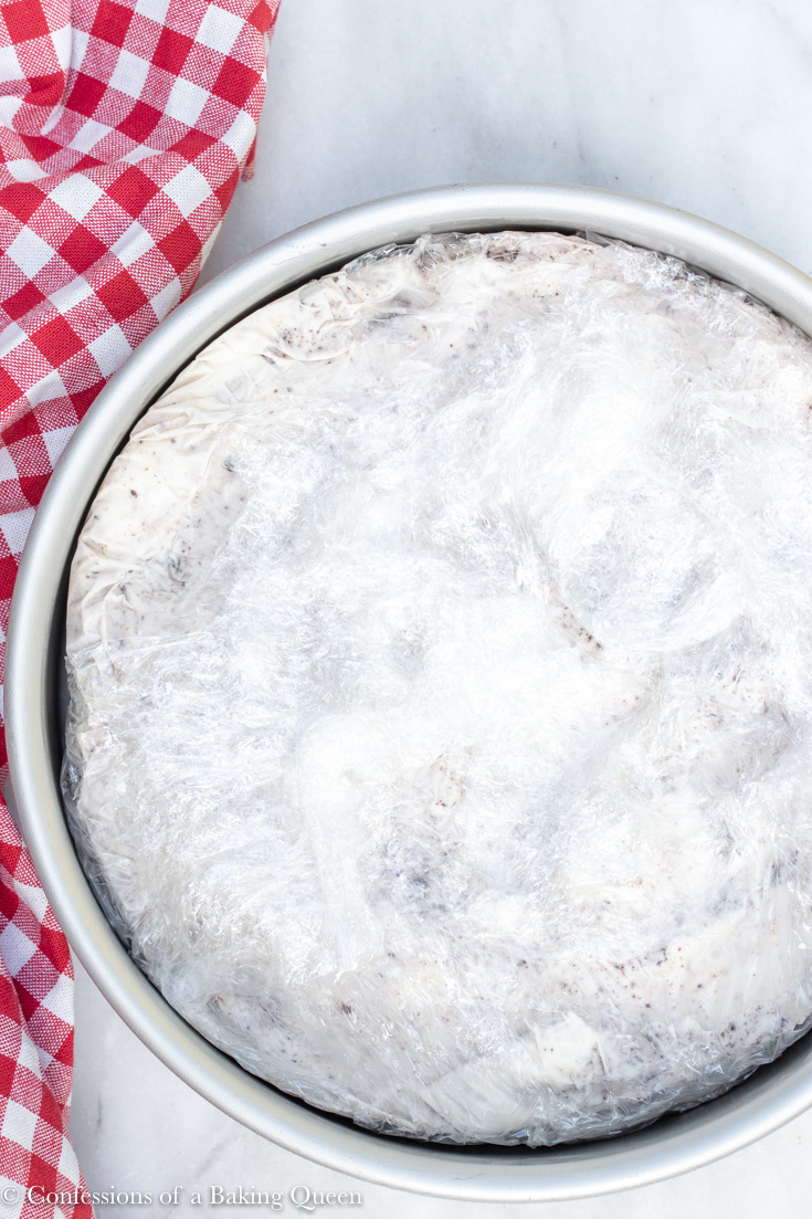 ice cream cake layer wrapped in plastic wrap sitting in a silver pan next to a white and red checkered linen on a white surface