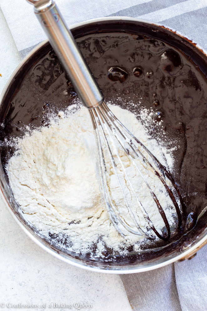 flour being added to the rest of ingredients for a peanut butter cup brownie recipe in a metal pot with a metal whisk sitting on a light surface with a grey linen