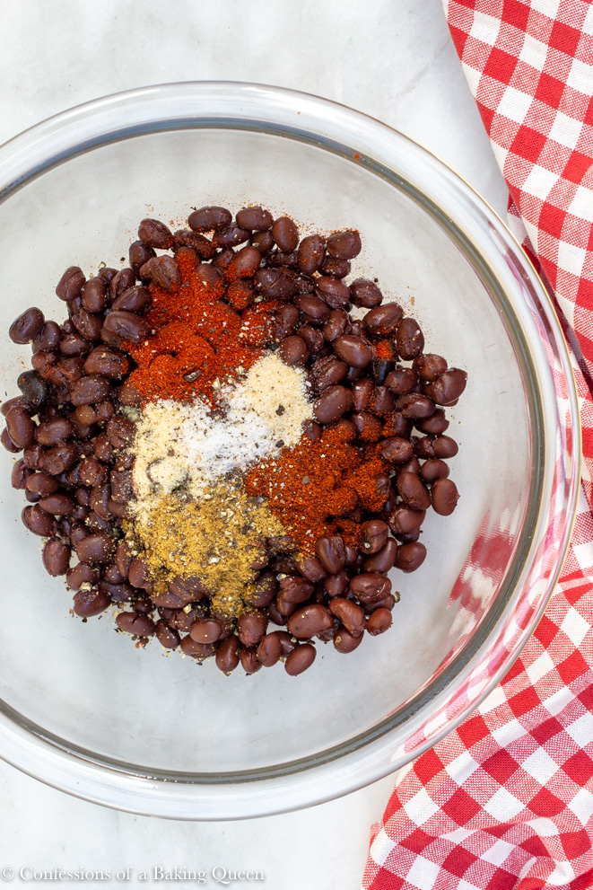 black beans and spices in a clear bowl on a white counter with a red and white checkered towel