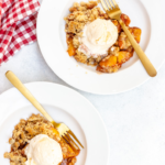 two white bowls full of a peach crumble recipe and vanilla ice cream with a red and white checkered towel off to the side