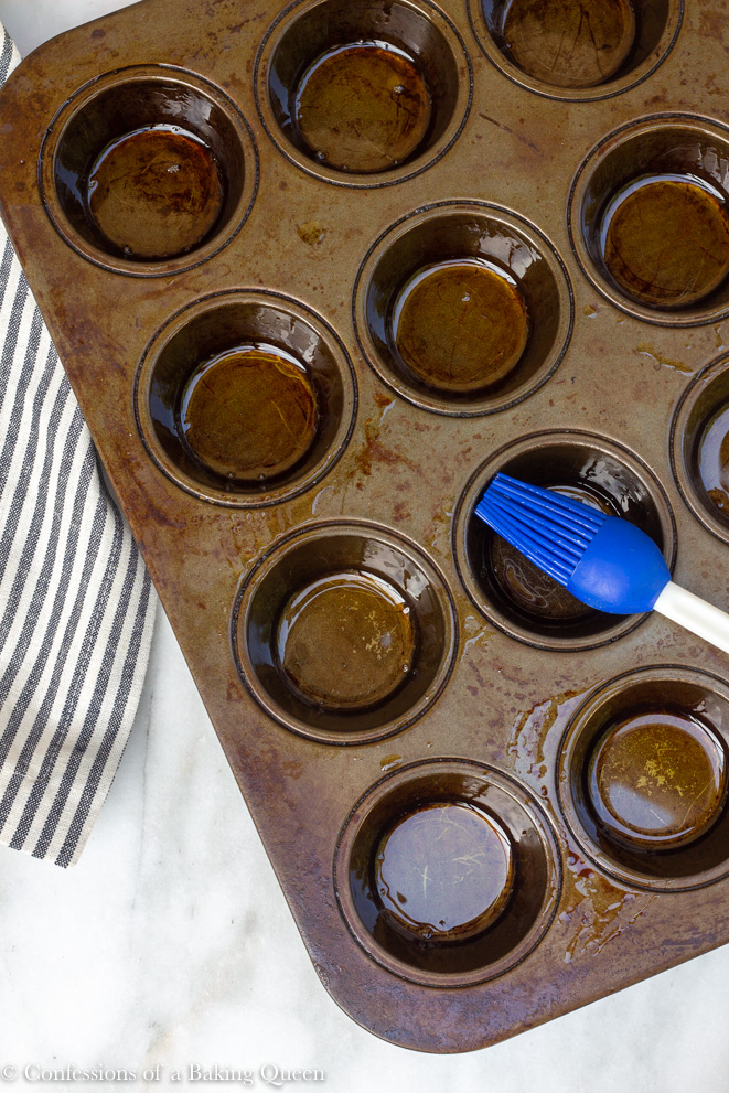 blue pastry brush brushing oil in a dark muffin tin on a white surface with a white and grey linen