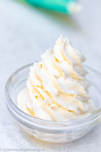 up close of Stabilized Whipped Cream in a clear bowl on a white surface with a piping bag in the background