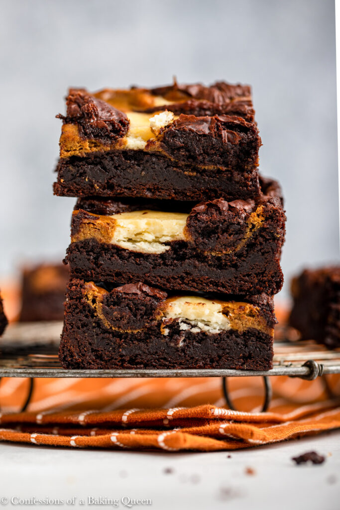 stack of pumpkin cream cheese brownies on a wire rack on top of an orange linen on a light grey surface
