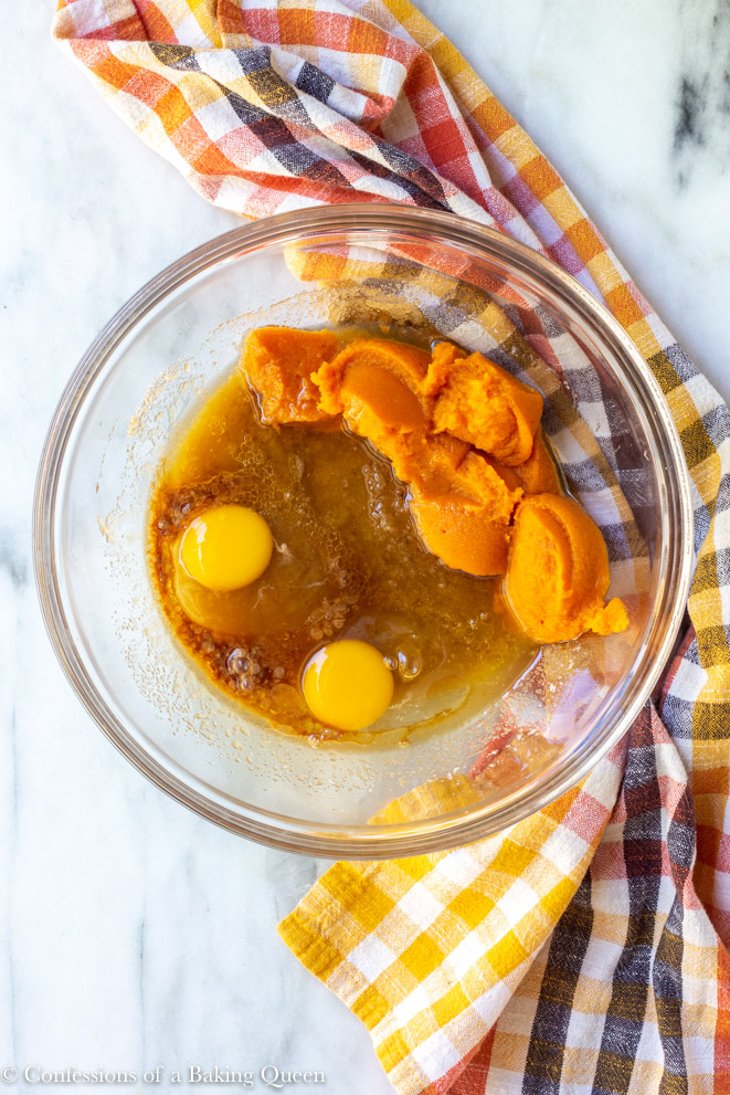 eggs and pumpkin puree added to butter sugar mixture in a clear bowl on a white surface with an orange, black, white and yellow linen