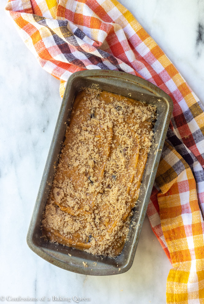 chocolate chunk pumpkin bread batter in a loaf pan with brown sugar sprinkled on top on a white surface with a yellow, orange, white, and black linen