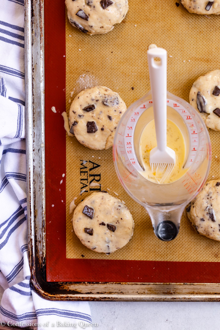 a measuring cup with an egg mixture and pastry brush sitting on a sheet pan of chocolate chunk scones before baking on a white surface with a white and blue linen 