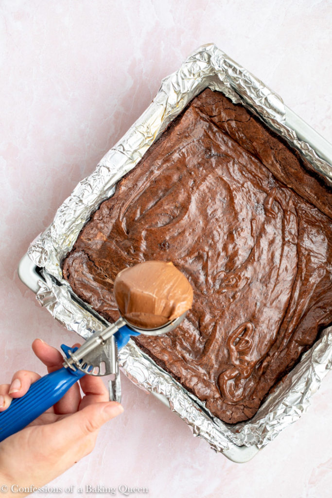 hand holding cookie scoop full of chocolate cheesecake batter to place on top of brownies