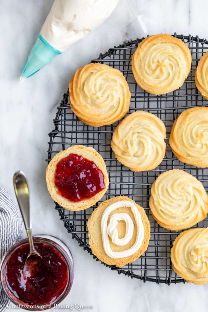 strawberry jam and a pastry bag with buttercream next to a wire rack of viennesse whirl biscuits on a white background with a white and blue linen