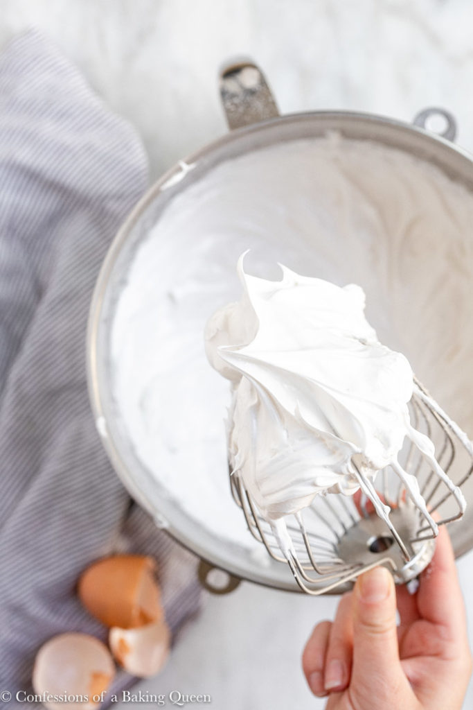 hand holding whisk of meringue cookies batter above a metal bowl on a marble surface with a white and blue linen
