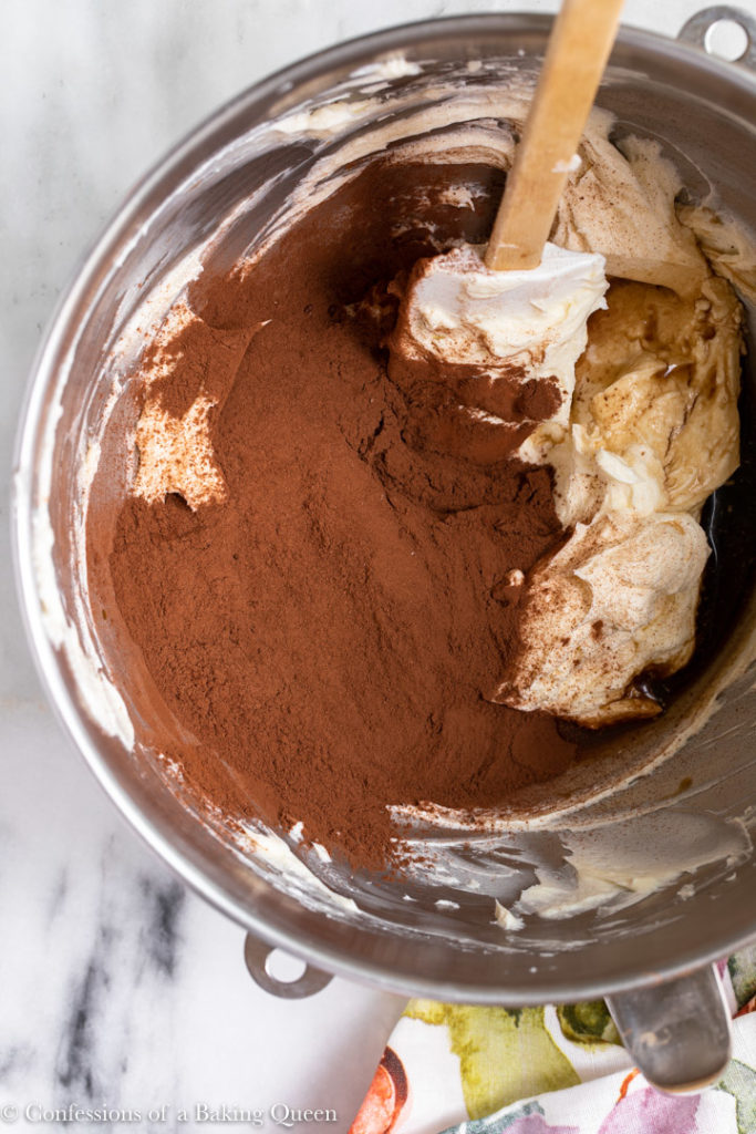 cocoa powder added to cream cheese mixture in a metal mixing bowl on a white marble surface with a floral linen