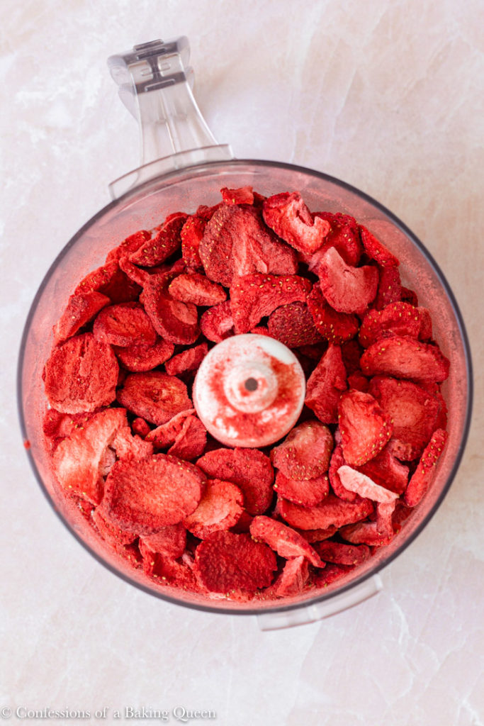 freeze dried strawberries in the bowl of a food processor on a light pink background
