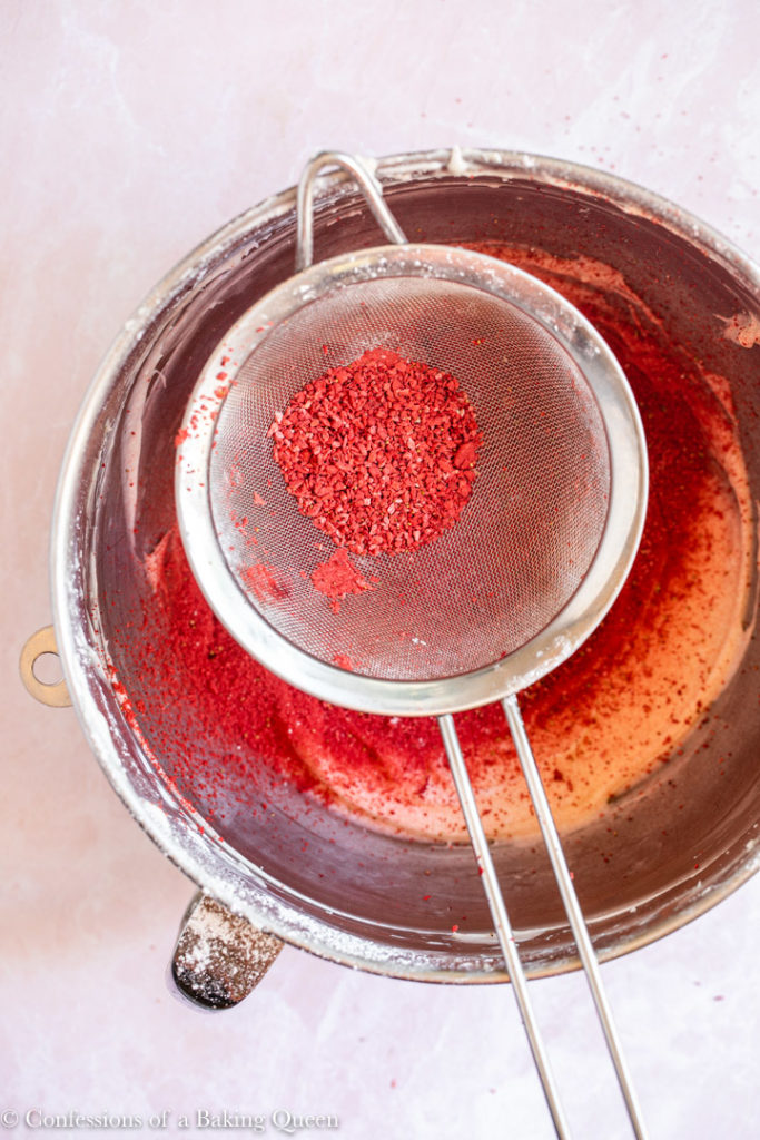 freeze dried strawberry in a metal sieve over frosting bowl on a light pink background