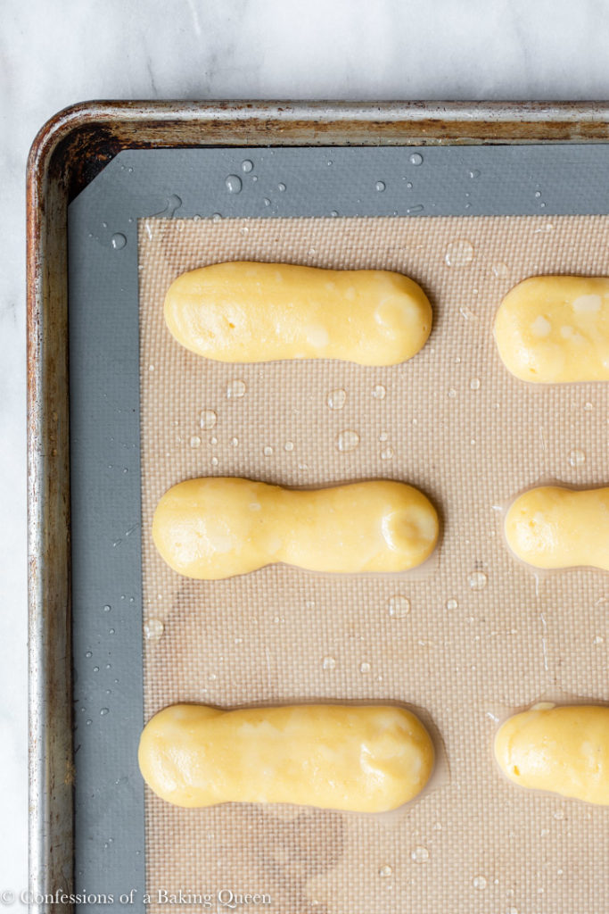 choux pastry piped out into an eclair shape on silpat lined baking sheet on a white marble surface