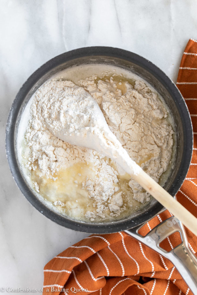 flour added to wet ingredients with a wooden spoon in a small saucepan on a white marble surface with an orange linen