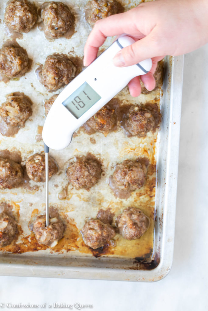 instant-read thermometer checking internal temperature of meatballs on a baking tray on a white marble surface 