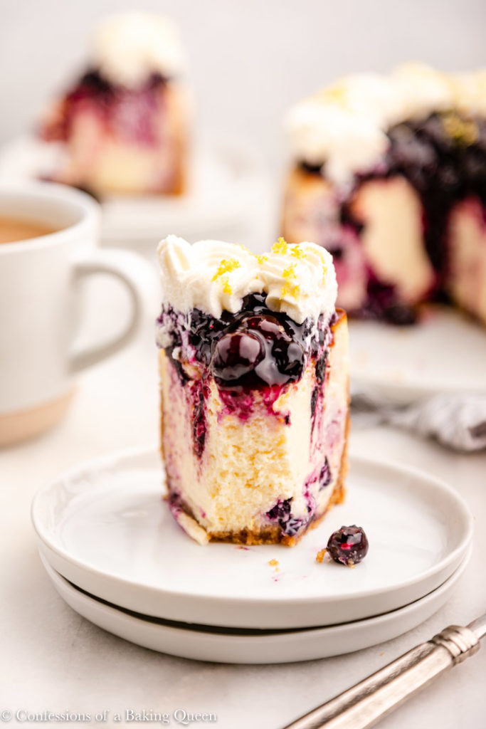 slice of lemon blueberry cheesecake on a white plate next to a cup of tea and fork and more cheesecake in the background all on a white surfacae