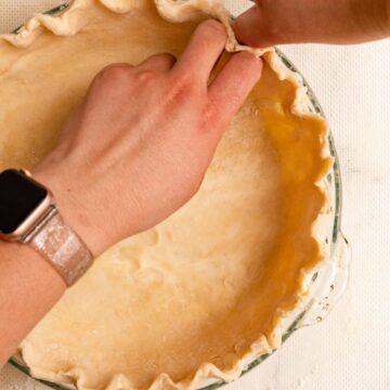 hand pressing pie dough into a crimped design in a glass pie plate on a white surface