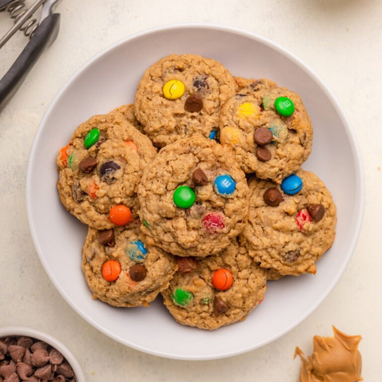 plate full of monster cookies next to a cup of chocolate chip, oats, and m&ms and a spoon full of peanut butter