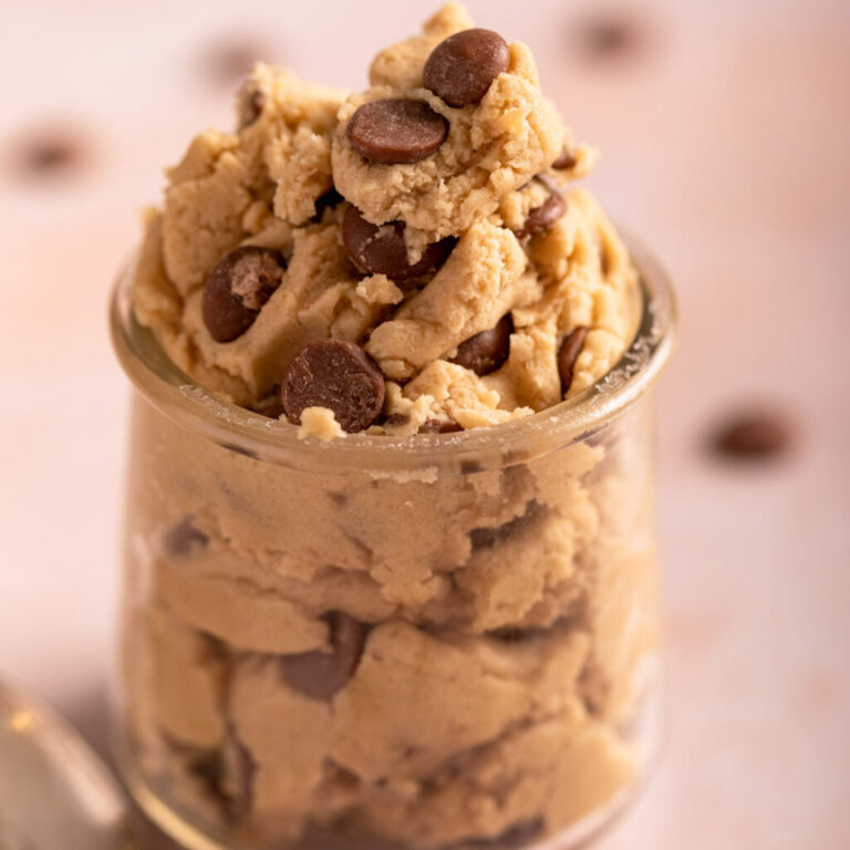 close up of edible chocolate chip cookie dough in a glass jar on a light pink surface with a spoon and extra chocolate chips