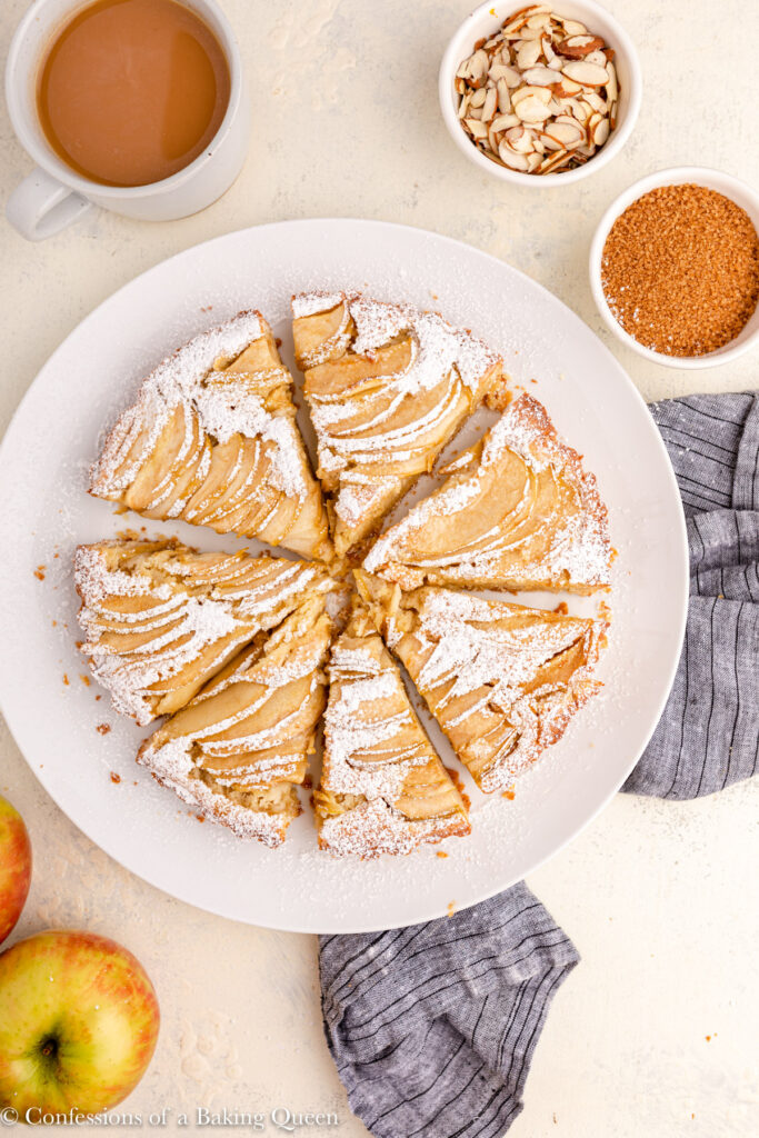 apple frangipane tart dusted with powdered sugar on a white plate next to a cup of coffee, apples, bowl of almonds and a bowl of sugar