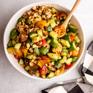 cucumber, corn, and tomato salad in a white bowl with a wooden spoon next to a black and white checkered towel