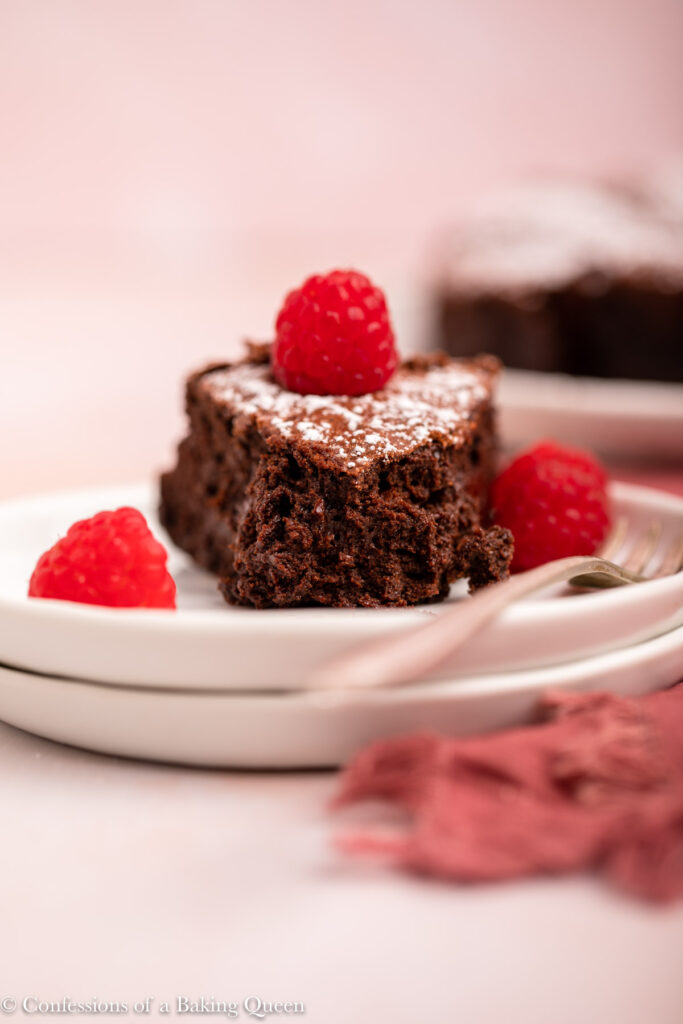 flourless chocolate cake on a white plate with raspberries and a fork with another cake in the background