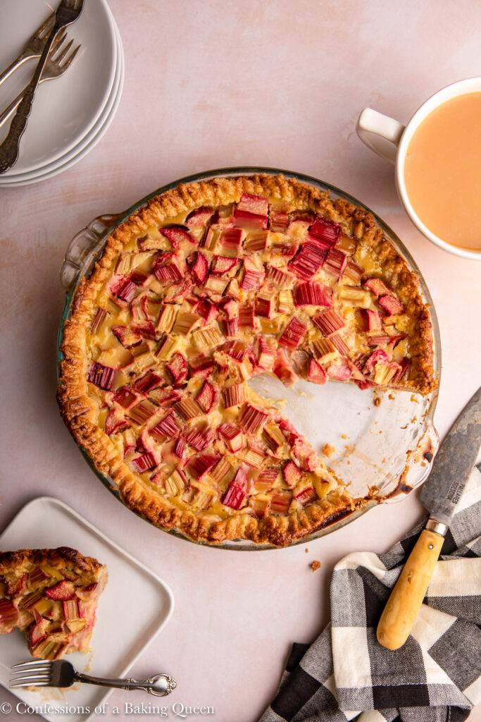 rhubarb custard pie missing a slice on a pink surface next to a slice of pie, cup of tea, more plates and forks, and a black and white linen