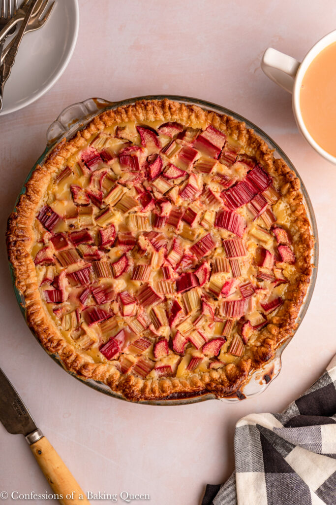 rhubarb custard pie on a pink surface next to a cup of tea, knife, black and white linen and plates with forks