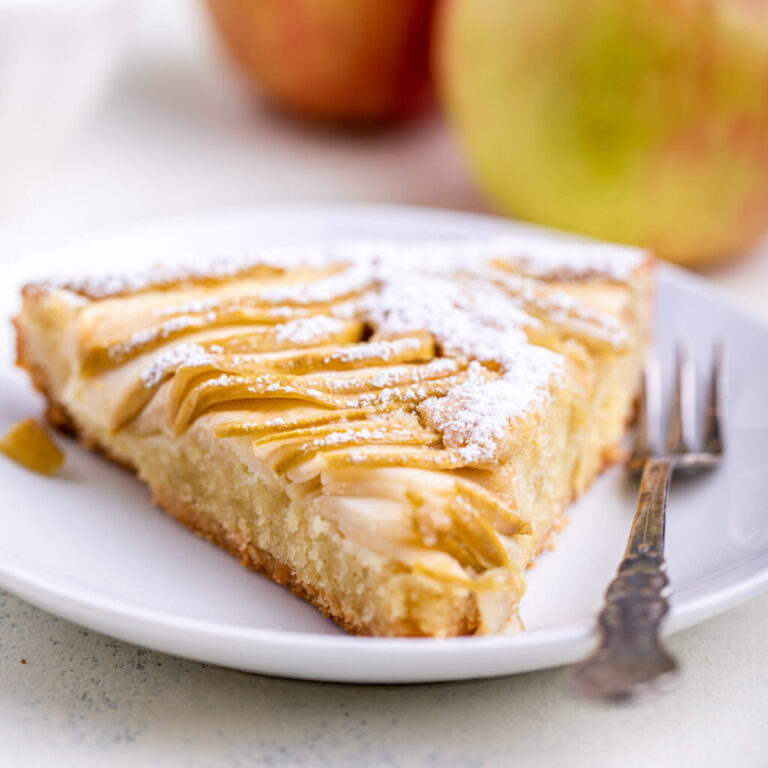slice of apple almond tart with a fork on a white plate with apples in the background