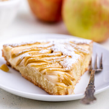 slice of apple almond tart with a fork on a white plate with apples in the background