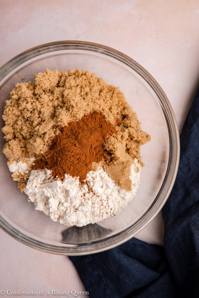 brown sugar, flour, salt, and ground cinnamon in a glass bowl on a pink surface next to a navy blue linen
