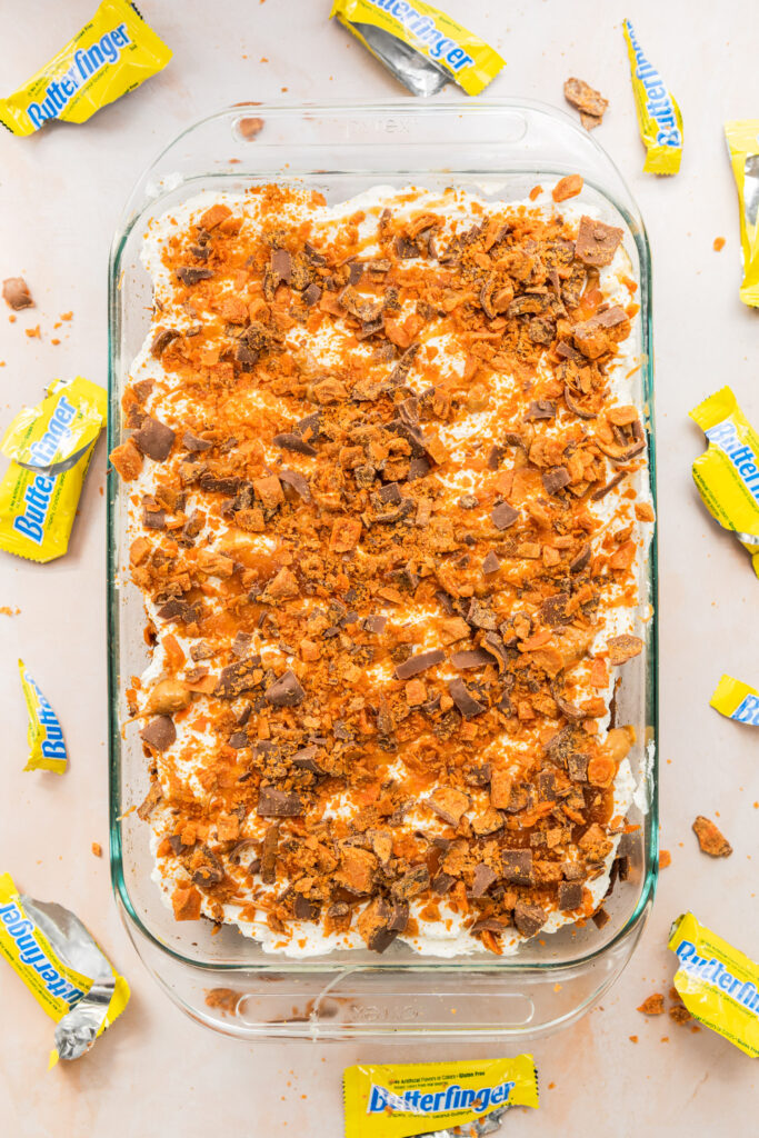 butterfinger poke cake in a glass dish on a pink surface with butterfinger candy wrappers all around