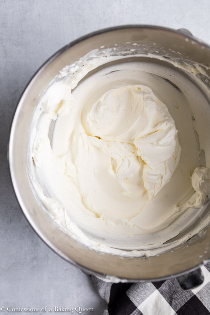 cream cheese whipped cream frosting- in a metal bowl with a wire whisk on a grey surface with a white and black linen