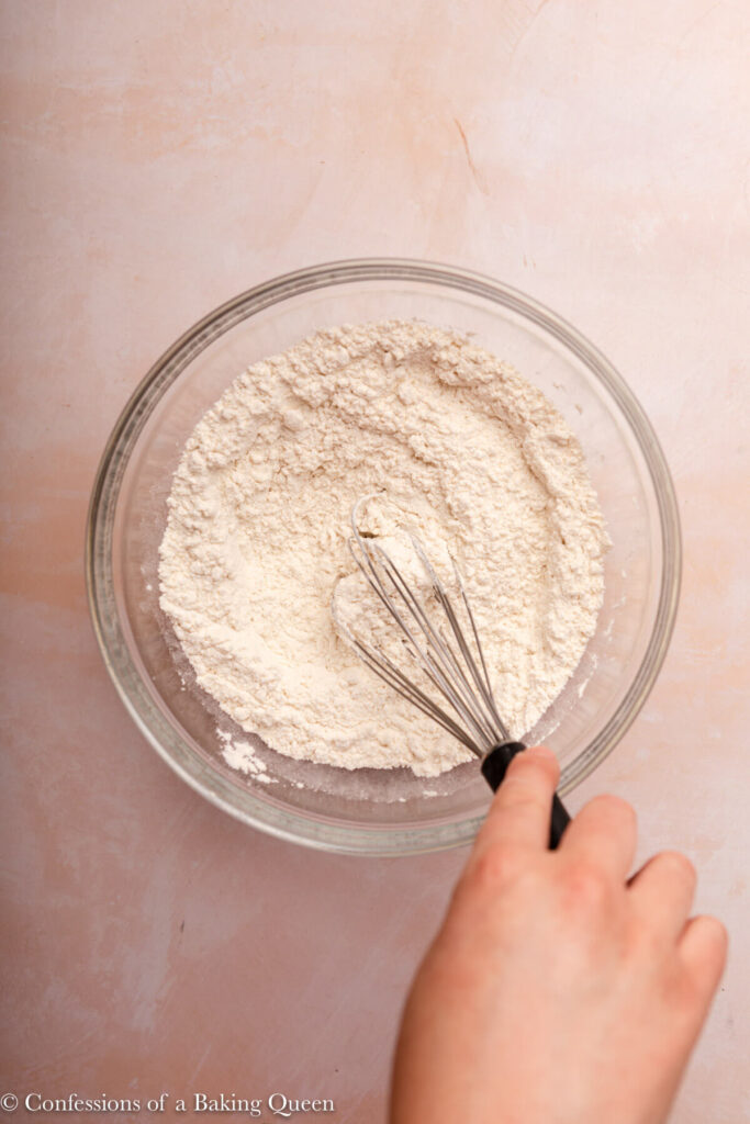 hand holding a whisk whisking dry ingredients in a glass bowl on a pink surface