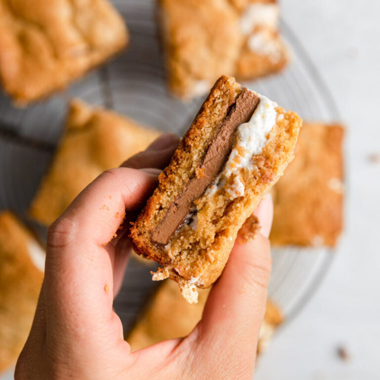hand holding smores bar to show inside filling on top of more bars on a wire rack on a grey surface