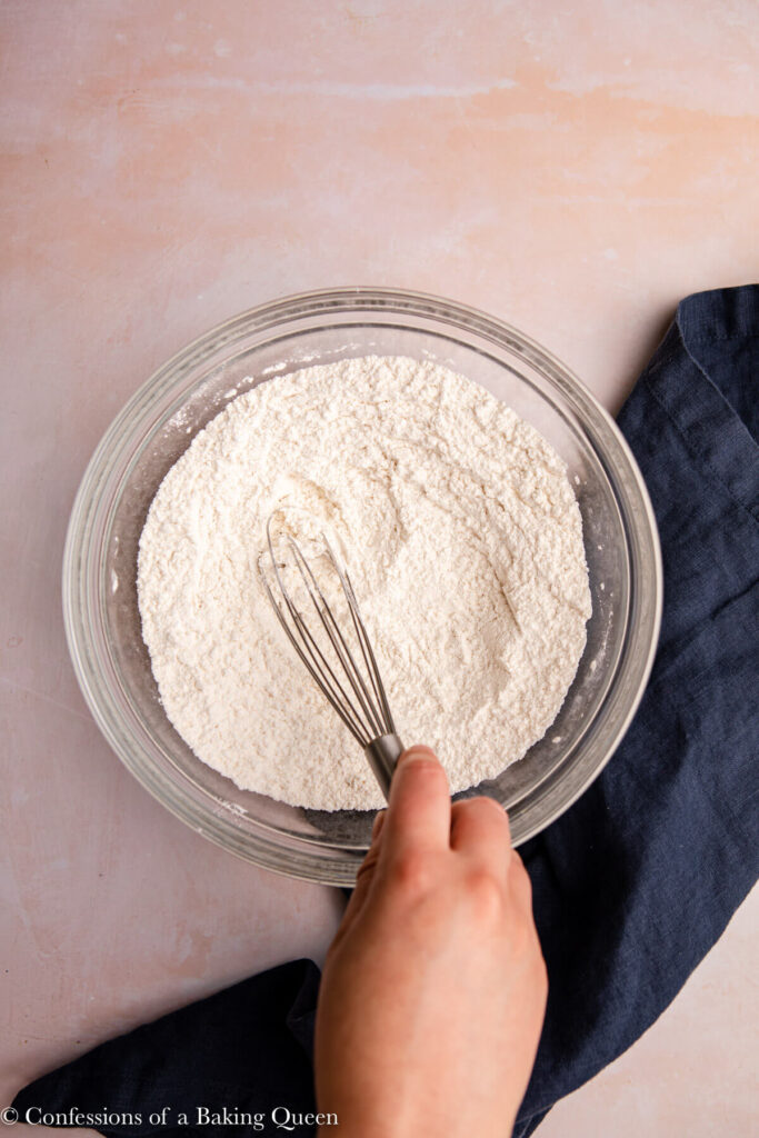 hand whisking dry ingredients in a glass bowl on a pink surface with a navy blue linen