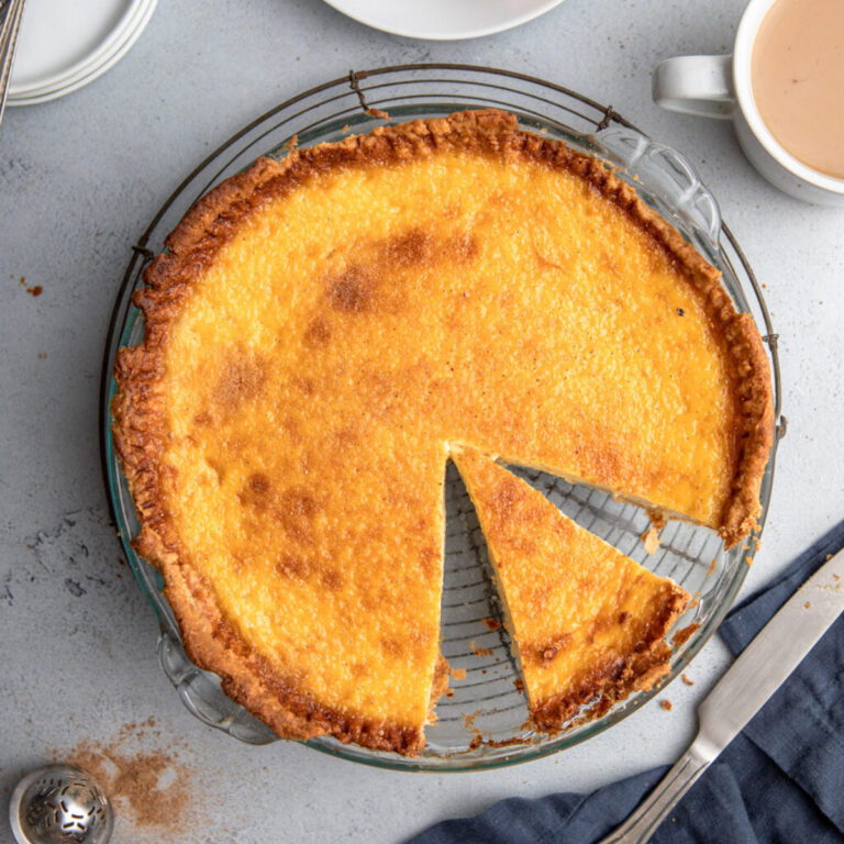cut open egg custard pie on a wire rack on a grey surface with a cup of tea, plates and forks, slice of pie on a white plate and a navy blue linen with a knife
