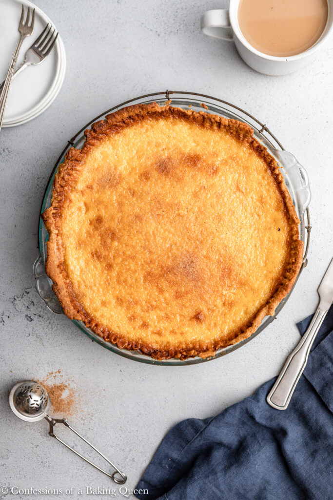 egg custard pie cooling on a wire rack with a cup of tea, plates and forks, and blue linen on a grey surface 