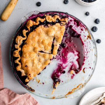 half eaten blueberry pie in a glass dish on a grey surface with a cup of milk, bowl of blueberries, knife, and slice of pie on a plate next to the pie plate