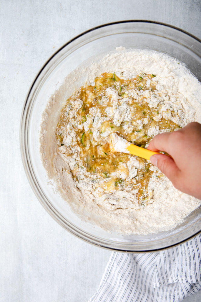hand folding dry ingredients into cake batter in a glass bowl on a grey surface with a white and blue linen