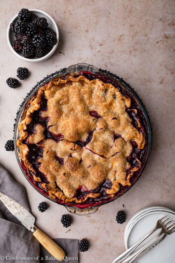 blackberry pie cooling on a wire rackon a light brown surface with a grey linen, slice of pie on a plate, and bowl of blackberries