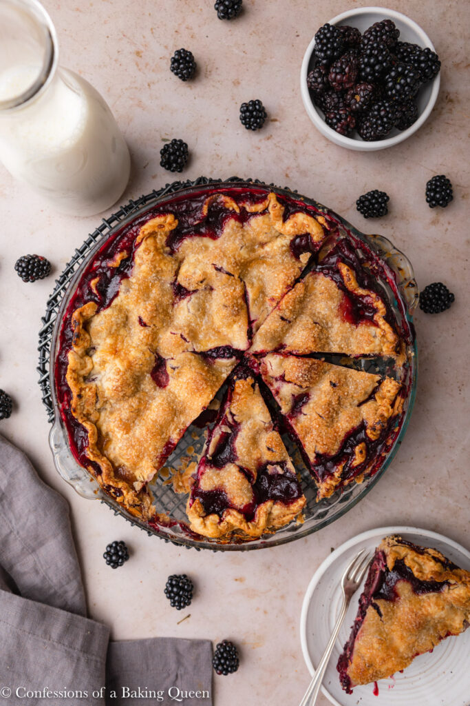 blackberry pie cut into slices on a light brown surface with a grey linen, slice of pie on a plate, bowl of blackberries and milk carafe