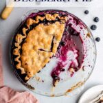 half eaten blueberry pie in a glass dish on a grey surface with a cup of milk, bowl of blueberries, knife, and slice of pie on a plate next to the pie plate