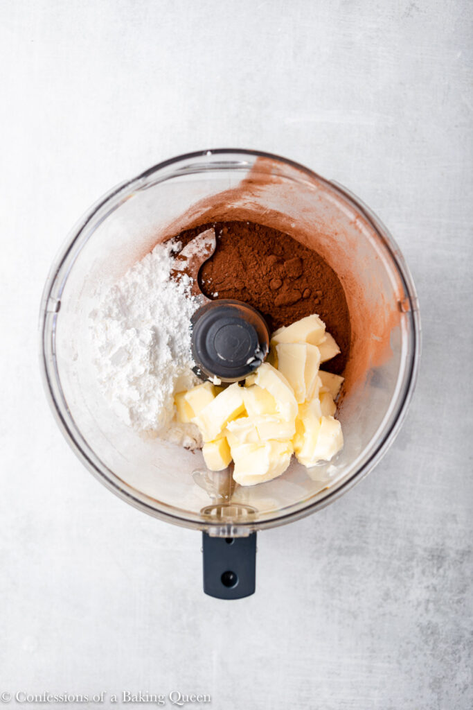 butter, powdered sugar, salt, and cocoa powder in the bowl of a food processor on a grey surface